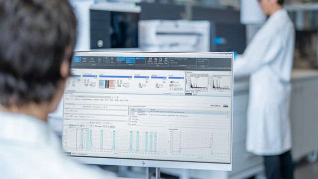 A scientist viewing analytical data on a computer screen with another scientist working at an instrument in the background. 