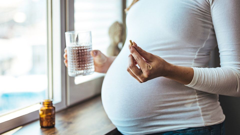 A pregnant person holding a pill in one hand and a glass of water in the other.