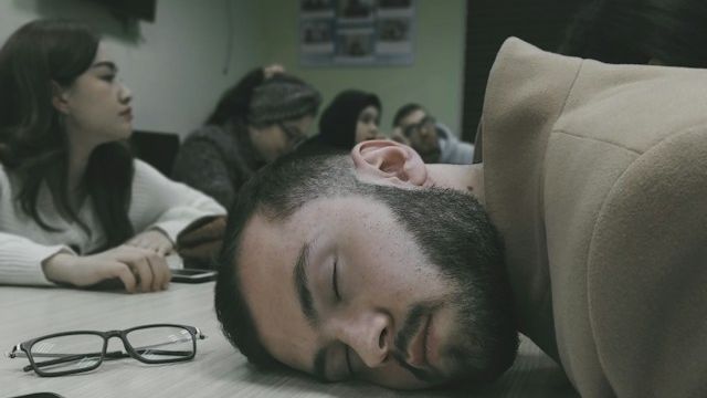 A person asleep with their on the desk with the other around are awake and chatting. 
